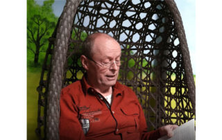 Eine lebensälterer Freiwilliger sitzt in einem Korbstuhl und liest etwas vor