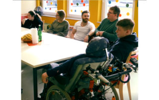 Sechs Personen, einer davon im Rollstuhl, sitzen um einen Tisch und unterhalten sich