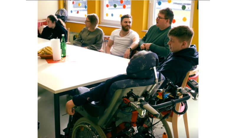 Sechs Personen, einer davon im Rollstuhl, sitzen um einen Tisch und unterhalten sich