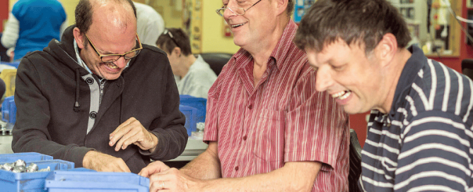 Ein älterer Freiwilliger arbeitet mit zwei zu Betreuenden in einer Werkstatt für Menschen mit Beeinträchtigung