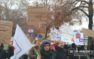 Ca. 10 Freiwillige bei einer Demonstration; sie heben Plakate in die Luft mit dem paritätischen Gleichheitszeichen und u.a. der Forderung "Freie Fahrt für Freiwillige"