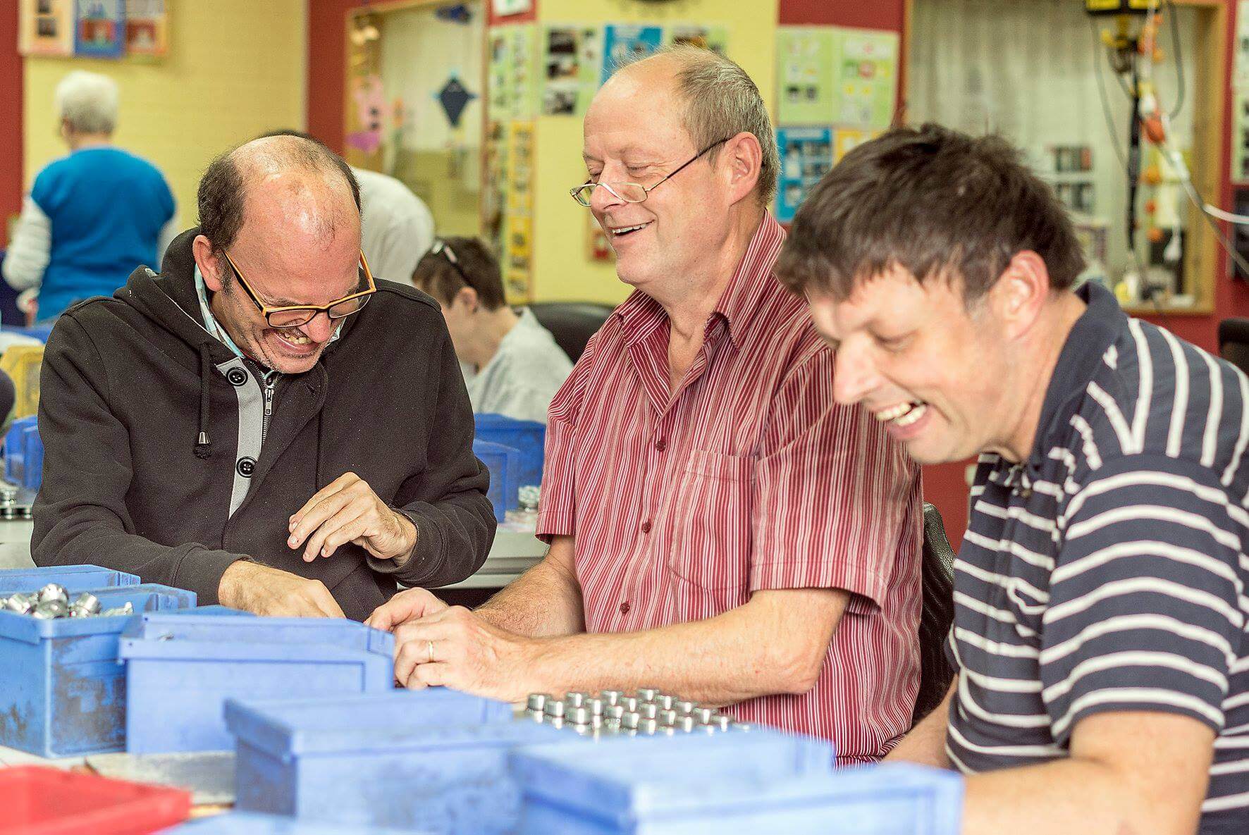 ein älterer Freiwilliger arbeitet mit zwei Klienten in einer Werkstatt für Menschen mit Beeinträchtigung