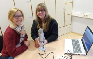 Annika Schäfer und Ina Rehor sitzen an einem Tisch mit Mikrofon und Laptop und lächeln in die Kamera
