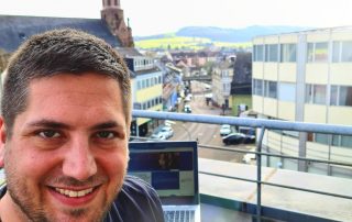 Kai Silbermann lacht von einer Dachterrasse in die Kamera; im Hintergrund sind sein Laptop und Häuser sowie eine Kirche zu sehen.