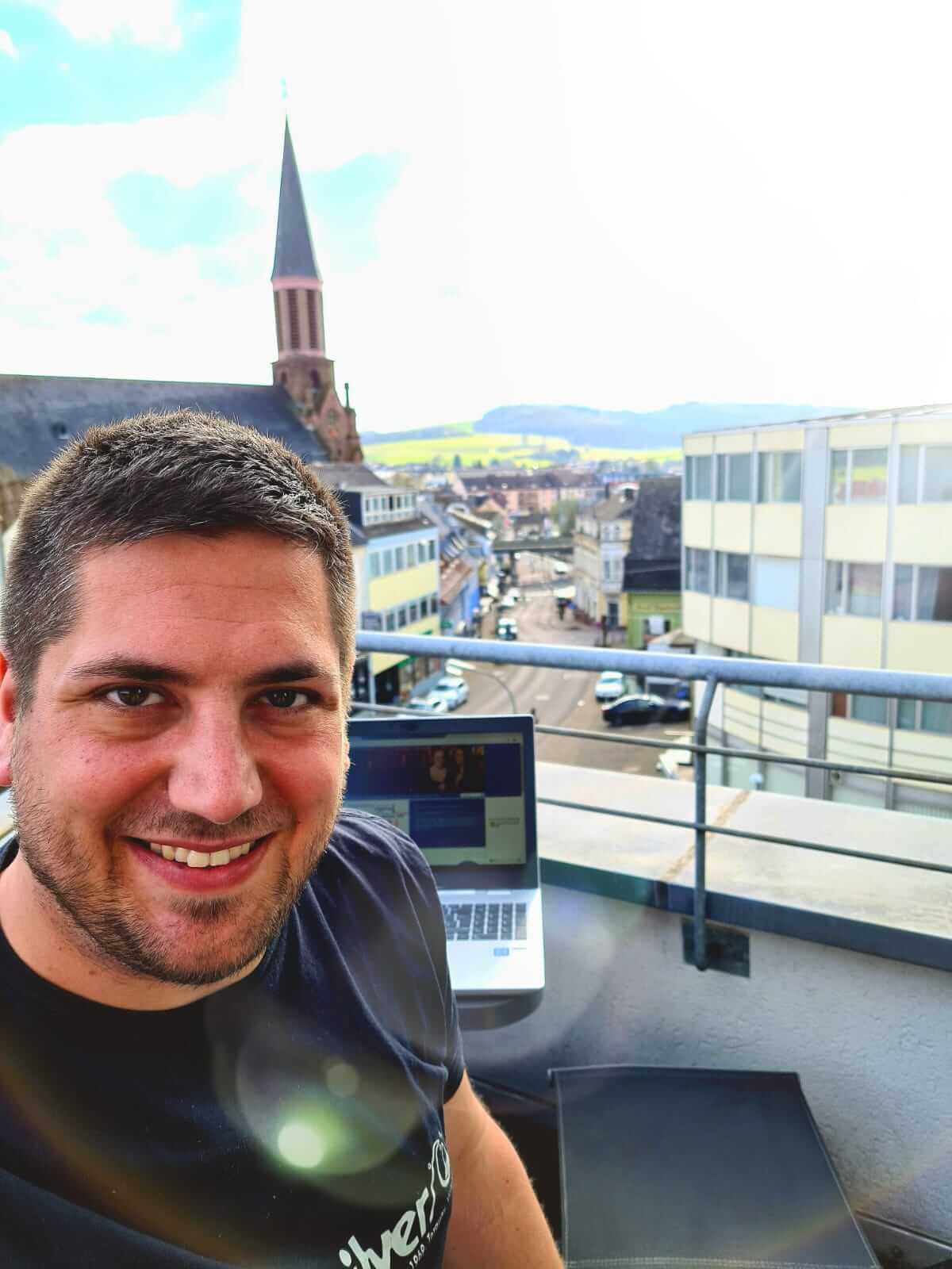 Kai Silbermann lacht von einer Dachterrasse in die Kamera; im Hintergrund sind sein Laptop und Häuser sowie eine Kirche zu sehen.