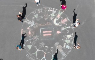 mehrere Menschen stehen um ein mit Kreide gemaltes Bild zum Motto gemeinsame Kreidezeit, sie wnken in die Kamera