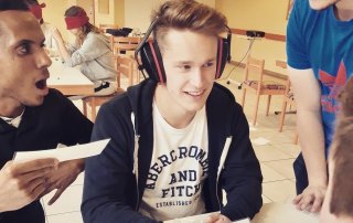 Mehrere Freiwillige sitzen an einem Tisch, einer davon hat Ohrenschützer an, während die anderem ihm etwas vorlesen. Im Hintergrund sitzen Freiwillige mit verbundenen Augen.