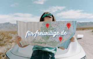 Frau sitzt auf der Motorhaube eines Autos und hält eine Karte in der Hand auf der "für-freiwillige.de" steht